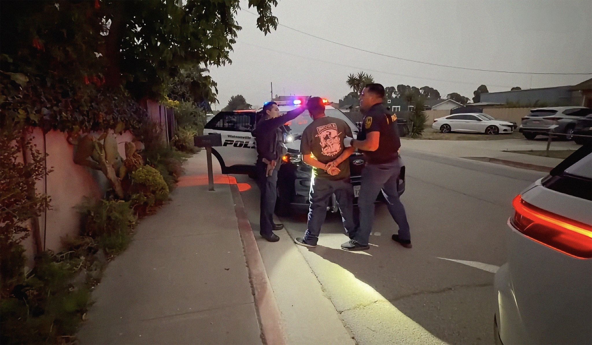 Two officers interact with a person near a police vehicle on a residential street at dusk.