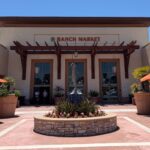 The Ranch Market storefront features a central fountain, potted plants, and orange umbrellas under a clear blue sky.