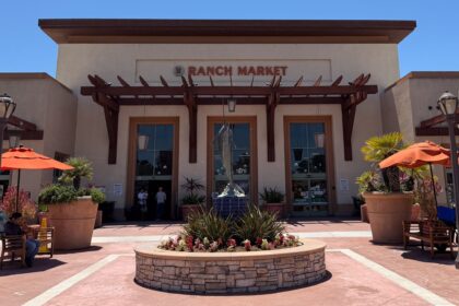 The Ranch Market storefront features a central fountain, potted plants, and orange umbrellas under a clear blue sky.