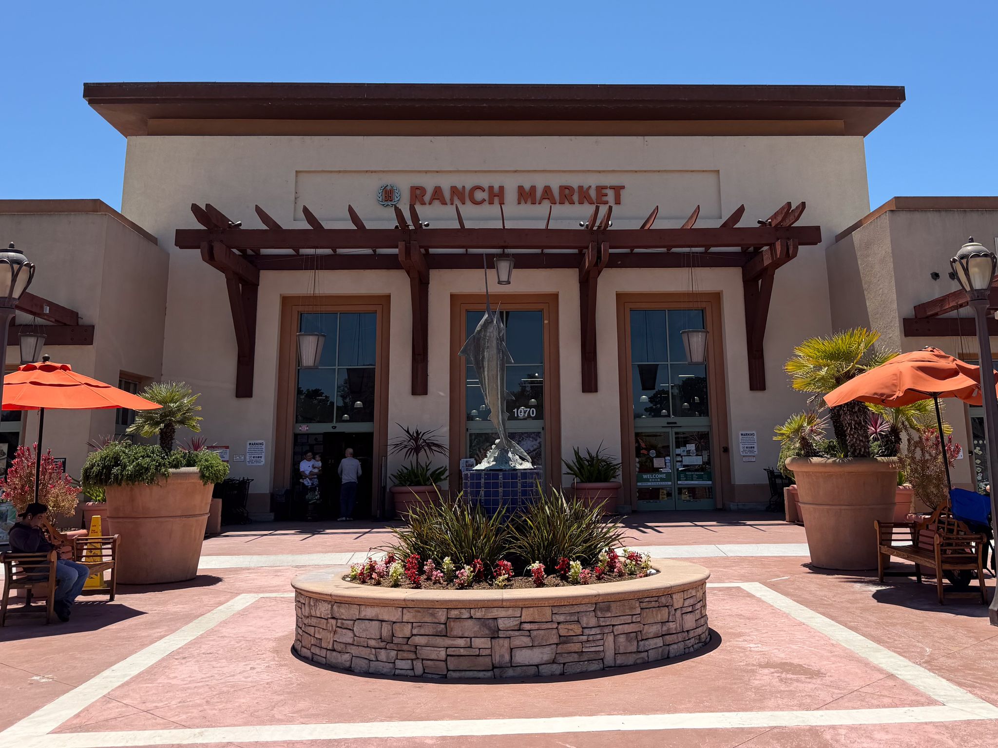 The Ranch Market storefront features a central fountain, potted plants, and orange umbrellas under a clear blue sky.